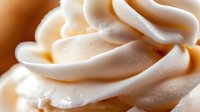 Creamy white whipped frosting swirls in macro shot showing soft peaks and smooth texture against warm background, fresh dairy dessert topping for cakes and cupcakes.
