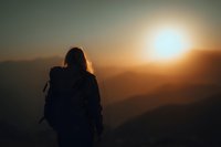 Silhouette of female hiker with backpack watching sunset over mountain ranges, golden light illuminating layered hills in warm atmospheric haze.