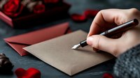 Hand writing romantic letter with fountain pen on kraft paper surrounded by red roses and envelope for Valentine's Day or anniversary celebration.