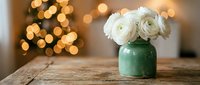 White ranunculus flowers in green ceramic vase on rustic wooden table with warm bokeh lights creating cozy home decor atmosphere for interior design.