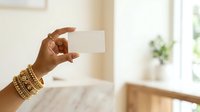 Young woman with medium skin tone wearing gold bracelets holds blank white business card in bright, modern interior space with natural window light.