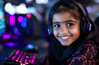 Young South Asian girl wearing hijab and gaming headphones smiles brightly while sitting at computer setup with colorful RGB lighting and gaming keyboard in background.