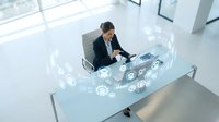 Asian male professional in dark business suit working at modern glass desk with holographic digital interface displaying technology icons and data visualization in bright office environment.