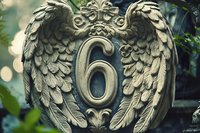 Ornate stone angel wings frame number six on decorative architectural element against blurred natural background with bokeh lights and foliage.