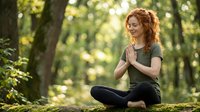 Young redhead woman practicing yoga meditation in peaceful forest setting with hands in prayer position sitting cross-legged on moss-covered log.