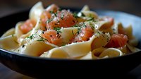 Fresh pappardelle pasta with smoked salmon pieces and dill garnish served in dark ceramic bowl, selective focus on tender pink fish and herbs.
