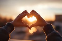 Hands forming heart shape against warm sunset sky creating romantic silhouette with golden light shining through fingers during golden hour outdoors.