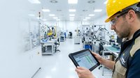 Male technician wearing yellow safety helmet and work uniform operating tablet in modern industrial manufacturing facility with machinery and equipment.