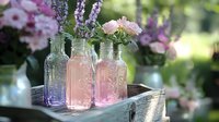 Vintage glass bottles with fresh flowers arranged on rustic wooden tray in garden setting. Pink roses and purple lavender create romantic summer atmosphere with soft natural lighting.