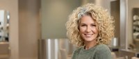 Smiling middle-aged Caucasian woman with curly blonde hair wearing green sweater in modern kitchen interior for lifestyle and home concepts.