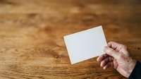 Close-up of mature hand holding blank white card against wooden surface, suitable for business branding, stationery mockups, and corporate identity presentations.