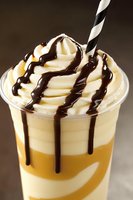 Creamy caramel frappuccino topped with whipped cream and chocolate sauce drizzle, served with striped straw against dark background.