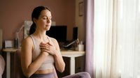 Young woman practicing mindfulness meditation at home, sitting peacefully with hand on chest in modern bedroom interior for wellness and mental health.