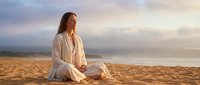 Young woman in white clothing meditating peacefully on sandy beach during golden hour sunset with soft cloudy sky creating serene wellness atmosphere.