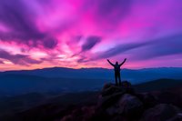 Silhouette of person with raised arms standing on mountain peak against dramatic purple and pink sunset sky over mountain ranges, expressing freedom and achievement.