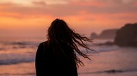 Young woman with long hair blowing in wind stands at ocean shore during vibrant sunset, silhouette against dramatic pink and orange sky.