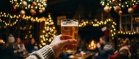 Hand holding a glass of beer at an outdoor winter celebration with golden string lights and festive decorations in the background.