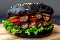 Juicy grilled meat chunks layered with fresh vegetables and lettuce between charcoal black burger buns, garnished with sesame seeds on wooden surface.