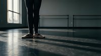 Dancer in black tights and pointe shoes standing at ballet barre in studio with natural light from window, professional training environment for classical dance instruction.