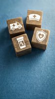 Four wooden blocks with engraved healthcare icons including ambulance, nurse cap, prescription bottle, and medical shield on blue fabric background.