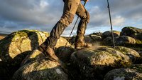 Hiker climbing rocky terrain with trekking poles, wearing rugged boots and outdoor pants, dramatic sky in the background.