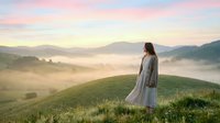 Woman in light cardigan standing on misty hillside at sunrise, overlooking fog-covered valley landscape with rolling mountains in soft morning light.