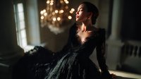 Young Hispanic woman in luxurious black lace evening gown poses elegantly near crystal chandelier in dimly lit classical interior, creating dramatic atmospheric portrait.
