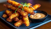 Crispy breaded chicken strips served on dark plate with creamy dipping sauce and microgreen garnish, close up view of golden-brown appetizer against wooden surface.