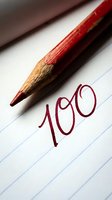 Red pencil writing number 100 on lined paper with close-up detail of pencil tip and handwritten text for education and academic achievement concepts.