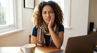 Young mixed-race woman with curly hair experiencing toothache pain at home office desk with laptop and coffee cup, holding cheek in discomfort.