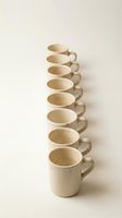 Eight handmade ceramic mugs arranged in diagonal line on neutral background showcasing artisan pottery craftsmanship and minimalist kitchen design aesthetic.