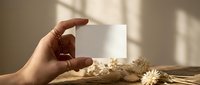Female hand holding blank white business card mockup with dried flowers and natural sunlight shadows on beige background for branding design.