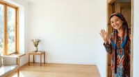 Woman with long dark hair and South Asian appearance wearing a colorful patterned blue tunic standing in doorway of bright minimalist home interior with wooden flooring, waving hello.