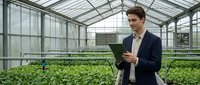 Young Caucasian businessman in suit using tablet in modern greenhouse with green plants for agricultural technology and smart farming solutions.