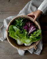 Fresh organic lettuce varieties and radicchio in woven basket on rustic wooden table with gray linen. Woman's hand holding basket of homegrown vegetables.