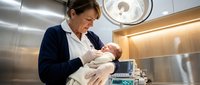 Caucasian middle-aged female nurse in scrubs gently holds a newborn baby in a modern hospital delivery room under surgical lights.