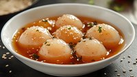 Traditional Asian sweet dumplings in amber syrup garnished with sesame seeds and herbs, served in white ceramic bowl for dessert menu photography.