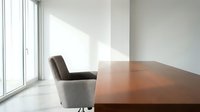 Modern minimalist office interior with wooden conference table and gray upholstered chair near large windows with natural sunlight streaming in.