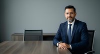 Mature male professional with dark hair and beard, wearing navy business suit and tie, seated at wooden conference table in modern office setting with gray walls and mesh chairs, displaying confident posture for corporate leadership and business concepts.