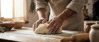 Senior baker kneading fresh dough on wooden table in rustic kitchen with flour and rolling pin. Traditional bread making process in warm sunlit bakery.
