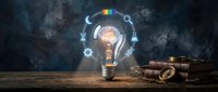 Glowing light bulb with brain inside surrounded by colorful icons on wooden desk with vintage books against dark smoky background for creative thinking.
