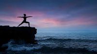 Young woman practicing warrior yoga pose on rocky cliff overlooking ocean at sunset with dramatic purple and pink sky for wellness and meditation concepts.