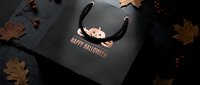 Black Halloween shopping bag with cauldron design surrounded by autumn leaves on dark surface for seasonal retail and holiday marketing campaigns.