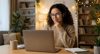 Young African American woman with curly hair and glasses working on laptop in cozy home office with warm lighting and holiday decorations in background.