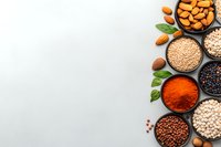 Assorted legumes, nuts, and spices in black bowls arranged on light gray background with copy space. Fresh basil leaves add natural accent.