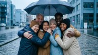 Diverse group of young adults huddling together under umbrella on rainy city street, smiling and embracing in friendship moment during urban weather.