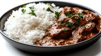 Savory beef stew with tender meat chunks in rich brown sauce served alongside fluffy white rice garnished with fresh parsley in black bowl.