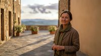 Senior woman enjoying peaceful moment on Mediterranean terrace during golden hour sunset with mountain views and historic stone architecture.