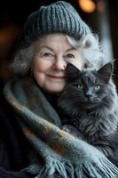 Senior Caucasian woman with silver hair wearing winter knit hat and scarf holds fluffy grey cat, both looking at camera with warm expressions in soft lighting.
