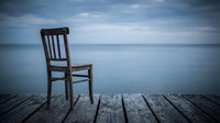 Wooden chair sitting alone on weathered dock overlooking calm ocean water and cloudy sky, conveying solitude, contemplation, and peaceful isolation for editorial and lifestyle use.